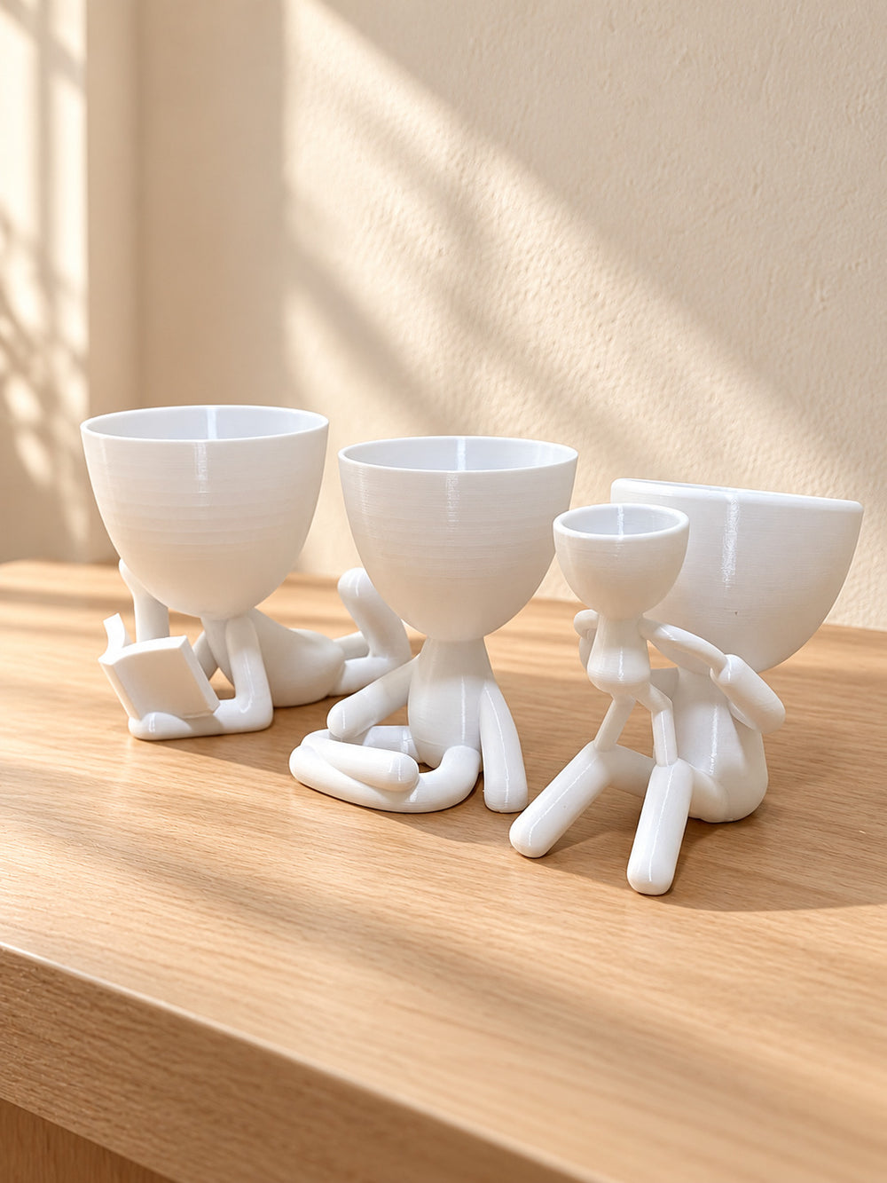 Três vasos brancos em forma de bonequinhos sentados sobre madeira, com sol na parede